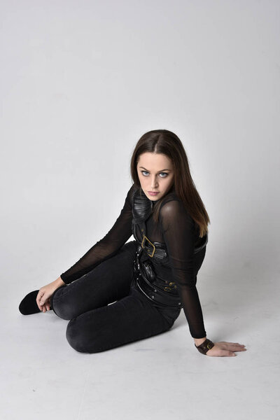 full length portrait of a pretty brunette woman wearing black leather fantasy costume. Crouching pose on a studio background.
