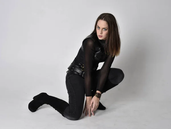 full length portrait of a pretty brunette woman wearing black leather fantasy costume. Crouching pose on a studio background.