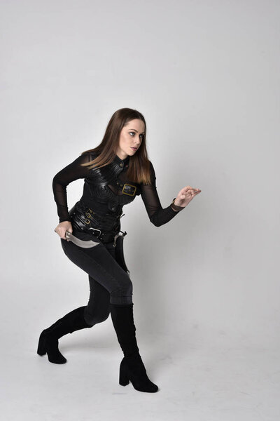 full length portrait of a pretty brunette woman wearing black leather fantasy costume. Standing pose, holding a dagger on a studio background.