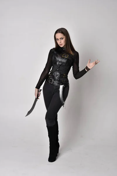 一个穿着黑色皮革幻想服装的漂亮的黑发女人的全长肖像 站立姿势 拿着匕首在工作室的背景上 — 图库照片