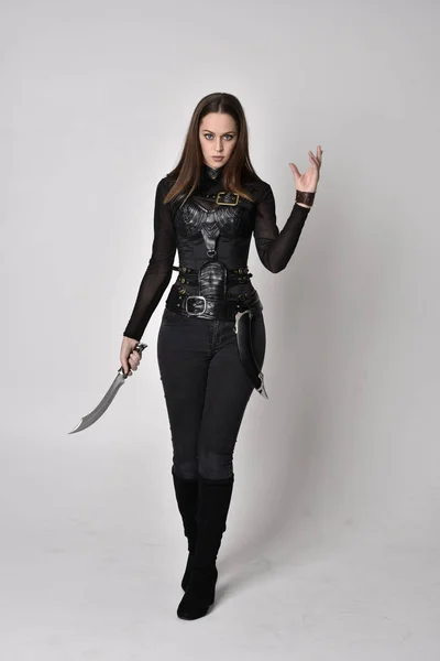 一个穿着黑色皮革幻想服装的漂亮的黑发女人的全长肖像 站立姿势 拿着匕首在工作室的背景上 — 图库照片