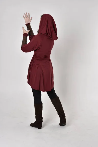 full length portrait of a girl wearing fantasy long red hooded tunic. standing pose with back to the camera.