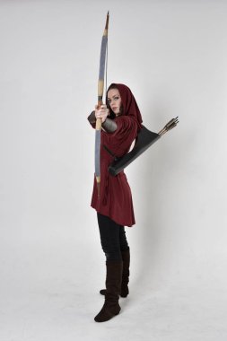 Kapüşonlu kırmızı fantezi bir ceket giyen esmer bir kızın uzun bir portresi. Elinde ok ve yay var. Beyaz bir stüdyonun arka planında poz vermek.