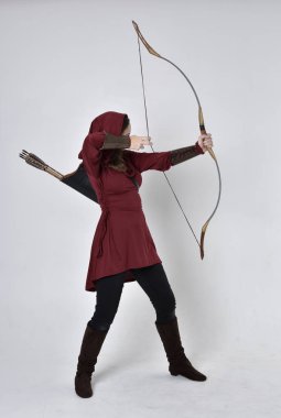 Kapüşonlu kırmızı fantezi bir ceket giyen esmer bir kızın uzun bir portresi. Elinde ok ve yay var. Beyaz bir stüdyonun arka planında poz vermek.