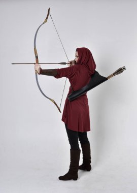 Kapüşonlu kırmızı fantezi bir ceket giyen esmer bir kızın uzun bir portresi. Elinde ok ve yay var. Beyaz bir stüdyonun arka planında poz vermek.