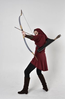 Kapüşonlu kırmızı fantezi bir ceket giyen esmer bir kızın uzun bir portresi. Elinde ok ve yay var. Beyaz bir stüdyonun arka planında poz vermek.