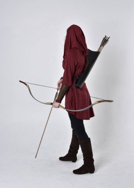 Kapüşonlu kırmızı fantezi bir ceket giyen esmer bir kızın uzun bir portresi. Elinde ok ve yay var. Beyaz bir stüdyonun arka planında poz vermek.
