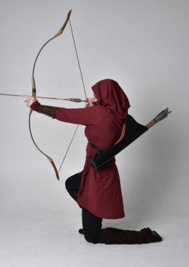 Kapüşonlu kırmızı fantezi bir ceket giyen esmer bir kızın uzun bir portresi. Elinde ok ve yay var. Beyaz bir stüdyonun arka planında otururken.