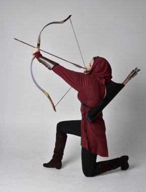 Kapüşonlu kırmızı fantezi bir ceket giyen esmer bir kızın uzun bir portresi. Elinde ok ve yay var. Beyaz bir stüdyonun arka planında otururken.