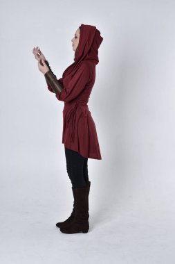 Kapüşonlu kırmızı fantezi ceket giyen esmer bir kızın tam boy portresi. Beyaz bir stüdyonun arka planında yan profilde duruyor..