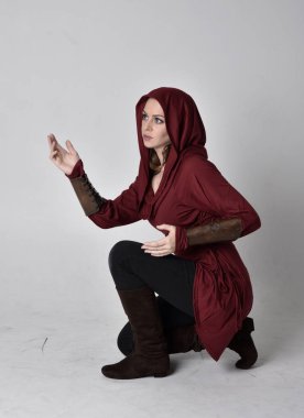 Kapüşonlu kırmızı fantezi ceket giyen esmer bir kızın tam boy portresi. Beyaz bir stüdyonun arka planında otururken.