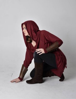 Kapüşonlu kırmızı fantezi ceket giyen esmer bir kızın tam boy portresi. Beyaz bir stüdyonun arka planında otururken.