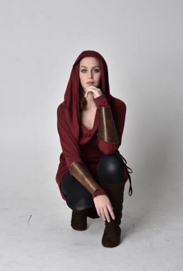 Kapüşonlu kırmızı fantezi ceket giyen esmer bir kızın tam boy portresi. Beyaz bir stüdyonun arka planında otururken.