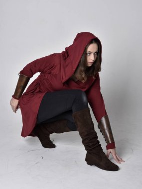 Kapüşonlu kırmızı fantezi ceket giyen esmer bir kızın tam boy portresi. Beyaz bir stüdyonun arka planında otururken.