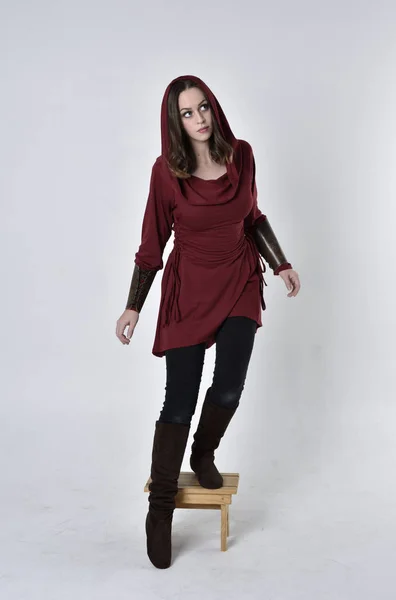 full length portrait of a brunette girl wearing a red fantasy tunic with hood. Stepping up standing pose on a white studio background.