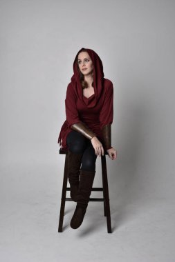 Kapüşonlu kırmızı fantezi ceket giyen esmer bir kızın tam boy portresi. Beyaz stüdyo geçmişi olan bir taburede oturuyor..