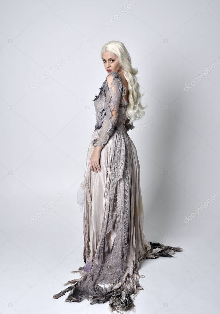 full length portrait of blonde girl wearing torn and ripped old bridal gown. standing pose with back to the camera  against a studio background.