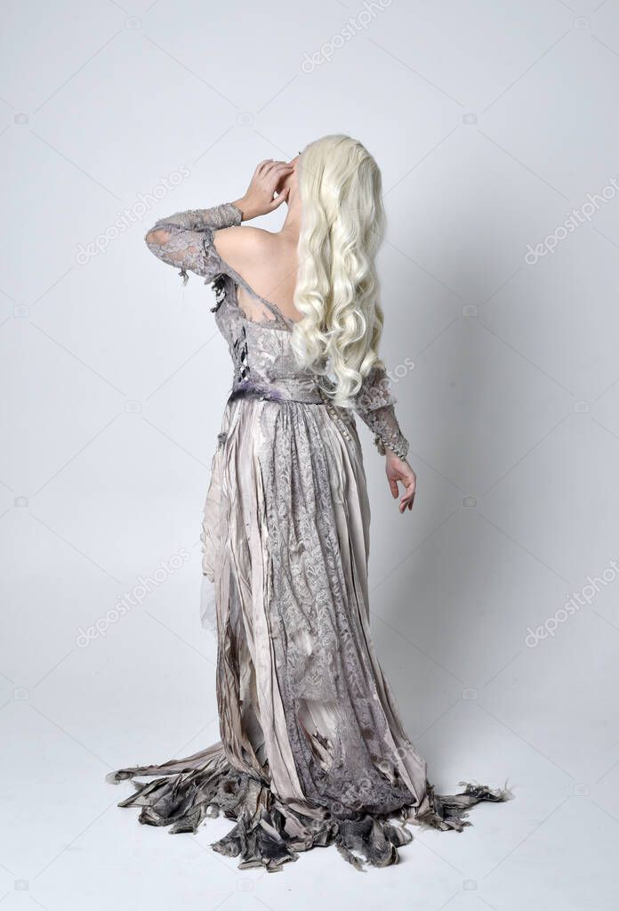full length portrait of blonde girl wearing torn and ripped old bridal gown. standing pose with back to the camera  against a studio background.