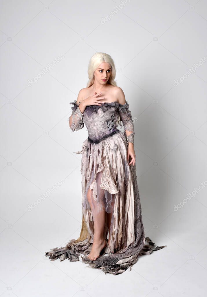 full length portrait of blonde girl wearing long torn old wedding dress.  standing pose with back to the camera on a studio background.