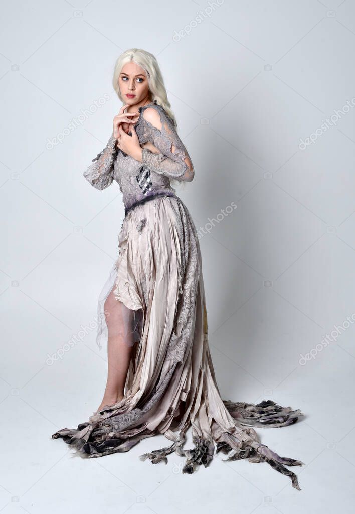 full length portrait of blonde girl wearing long torn old wedding dress.  standing pose with back to the camera on a studio background.