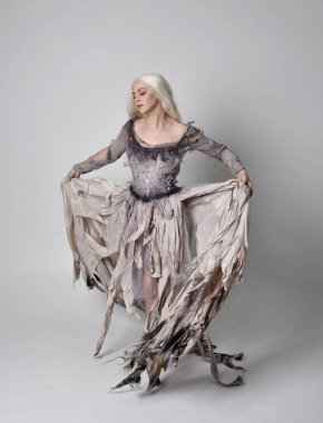 Uzun, yırtık, eski bir gelinlik giymiş sarışın kızın tam boy portresi. Stüdyonun arka planında kameraya dönük duruş.