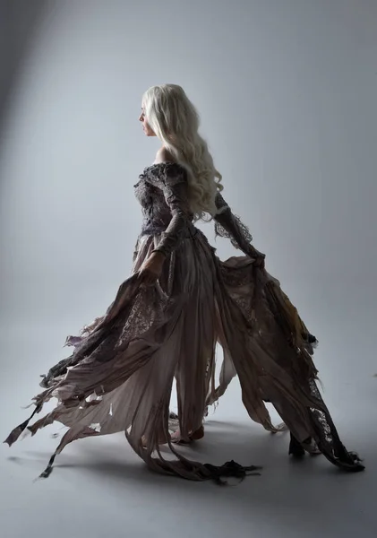 full length portrait of blonde woman wearing old torn wedding dress, standing pose with back to the camera on a grey studio background.