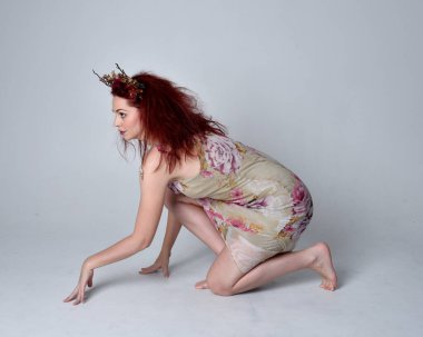 Çiçekli peri elbisesi ve başlık giyen kızıl saçlı kızın uzun pozu. gri stüdyo arka planında oturan poz.