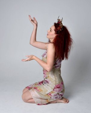 Çiçekli peri elbisesi ve başlık giyen kızıl saçlı kızın uzun pozu. gri stüdyo arka planında oturan poz.