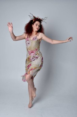 Çiçekli elbise ve başlık giyen kızıl saçlı kızın tam boy portresi. Gri stüdyo arka planında ayakta duruş.