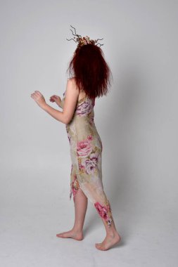 Çiçekli elbise ve başlık giyen kızıl saçlı kızın tam boy portresi. Gri stüdyo arka planında ayakta duruş.