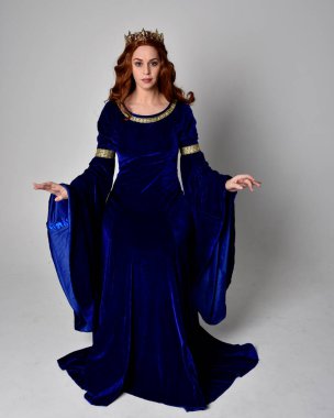 Altın taçlı, uzun mavi kadife bir elbise giyen kızın tam boy portresi. Oturma pozu, stüdyo arka planında izole..