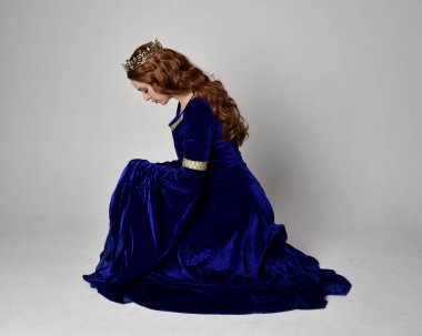 Altın taçlı, uzun mavi kadife bir elbise giyen kızın tam boy portresi. Oturma pozu, stüdyo arka planında izole..