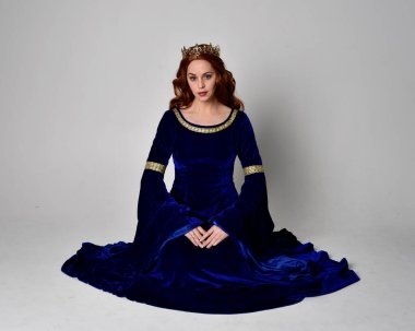 Altın taçlı, uzun mavi kadife bir elbise giyen kızın tam boy portresi. Oturma pozu, stüdyo arka planında izole..