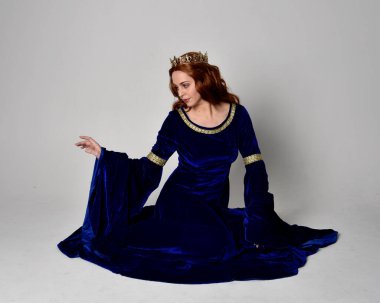 Altın taçlı, uzun mavi kadife bir elbise giyen kızın tam boy portresi. Oturma pozu, stüdyo arka planında izole..