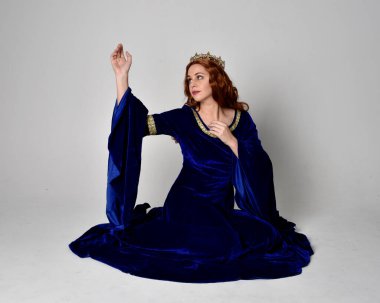 Altın taçlı, uzun mavi kadife bir elbise giyen kızın tam boy portresi. Oturma pozu, stüdyo arka planında izole..