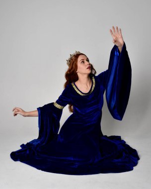 Altın taçlı, uzun mavi kadife bir elbise giyen kızın tam boy portresi. Oturma pozu, stüdyo arka planında izole..