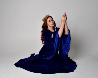 Altın taçlı, uzun mavi kadife bir elbise giyen kızın tam boy portresi. Oturma pozu, stüdyo arka planında izole..