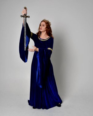 Uzun, mavi kadife elbisesi ve altın tacı olan, elinde kılıç tutan bir kızın portresi. Stüdyonun arka planında izole bir poz.