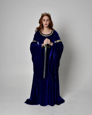Uzun, mavi kadife elbisesi ve altın tacı olan, elinde kılıç tutan bir kızın portresi. Stüdyonun arka planında izole bir poz.
