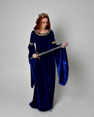 Uzun, mavi kadife elbisesi ve altın tacı olan, elinde kılıç tutan bir kızın portresi. Stüdyonun arka planında izole bir poz.