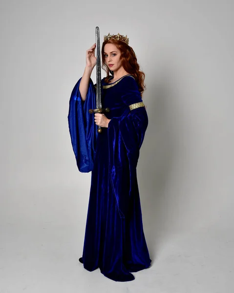 Full length portrait of  girl wearing long blue velvet gown with golden crown, holding a sword. standing pose  isolated against a studio background.