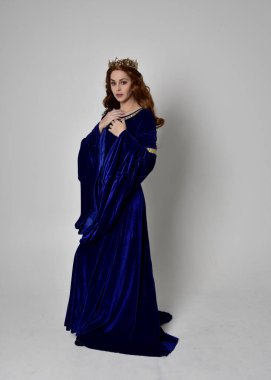 Altın taçlı, uzun mavi kadife bir elbise giyen kızın tam boy portresi. Kameraya dönük duruş, stüdyo arka planında izole edilmiş..