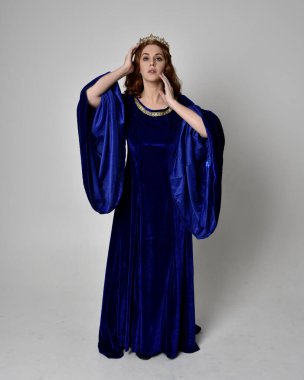 Altın taçlı, uzun mavi kadife bir elbise giyen kızın tam boy portresi. Kameraya dönük duruş, stüdyo arka planında izole edilmiş..