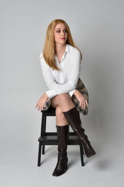 Beyaz gömlekli, ekose etekli ve çizmeli sarışın kızın tam boy portresi. Gri bir arka planda izole bir şekilde bir sandalyede oturuyor..