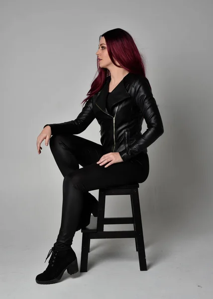 身穿黑色皮夹克 裤子和靴子的红头发女孩的全长肖像 坐在椅子上 与灰色的工作室背景隔离 — 图库照片