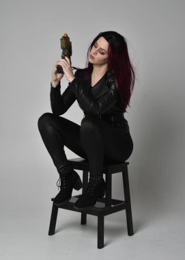 Kızıl saçlı, siyah deri ceketli, pantolonlu ve çizmeli bir kızın tam boy portresi. Elinde silah olan bir sandalyede oturan, gri bir stüdyonun arka planında izole edilmiş..
