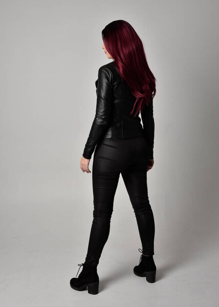 Full length portrait of  girl with red hair wearing black leather jacket, pants and boots. Standing pose facing away from the camera, isolated against a grey studio background.