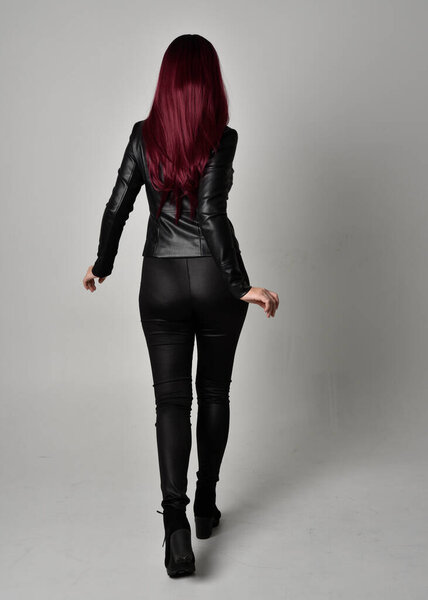 Full length portrait of  girl with red hair wearing black leather jacket, pants and boots. Standing pose facing away from the camera, isolated against a grey studio background.