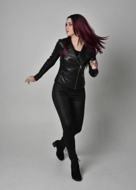 Kızıl saçlı, siyah deri ceketli, pantolonlu ve çizmeli bir kızın tam boy portresi. Gri bir stüdyonun arka planında tek başına poz veriyor..