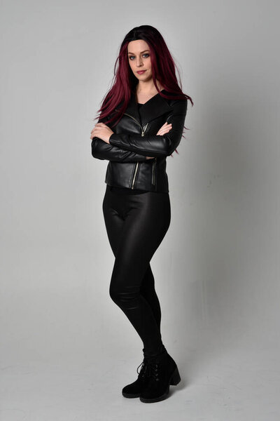 Full length portrait of  girl with red hair wearing black leather jacket, pants and boots. Standing pose, isolated against a grey studio background.